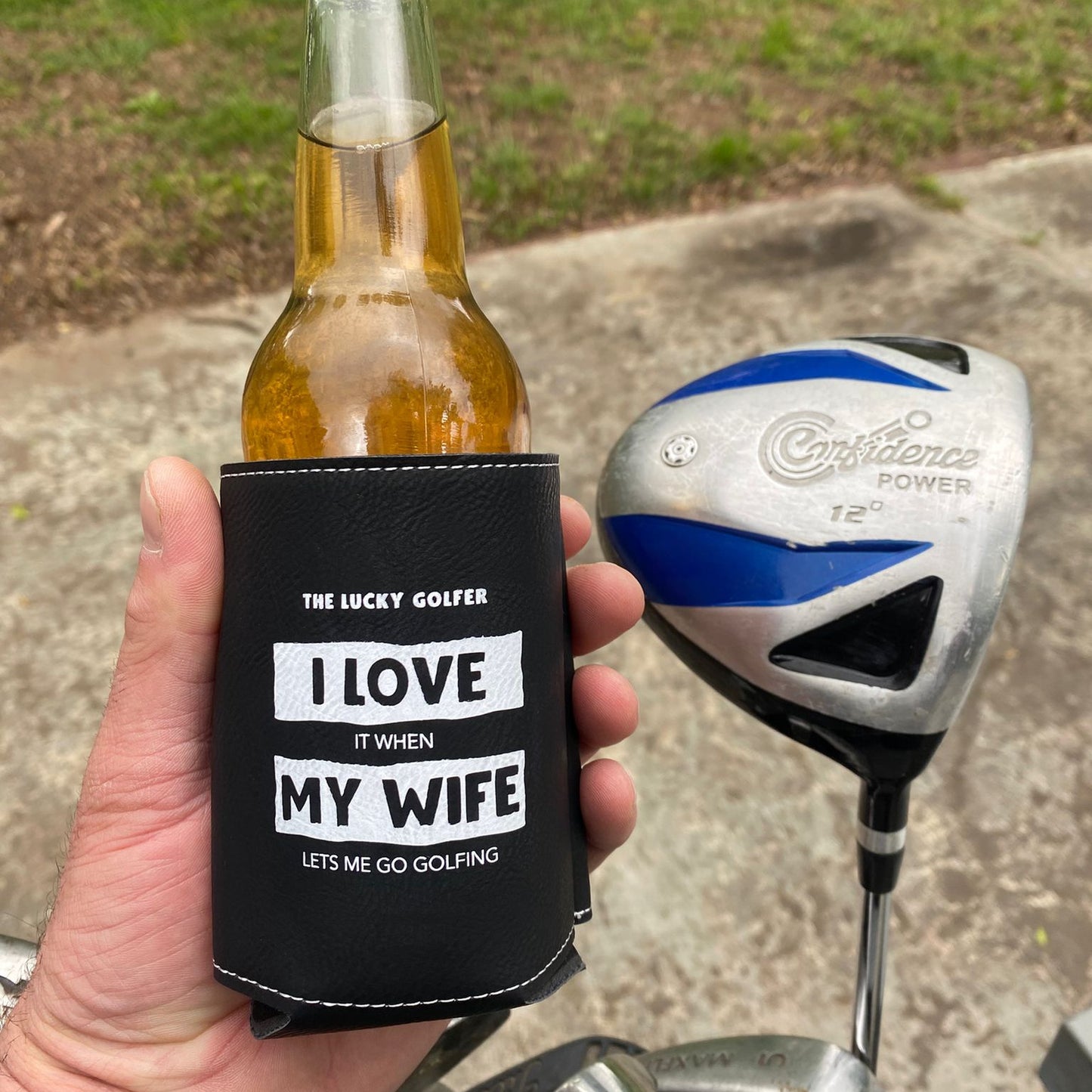 Hand holding a bottle with a can cooler labeled 'I Love My Wife' next to a golf club on a grassy background
