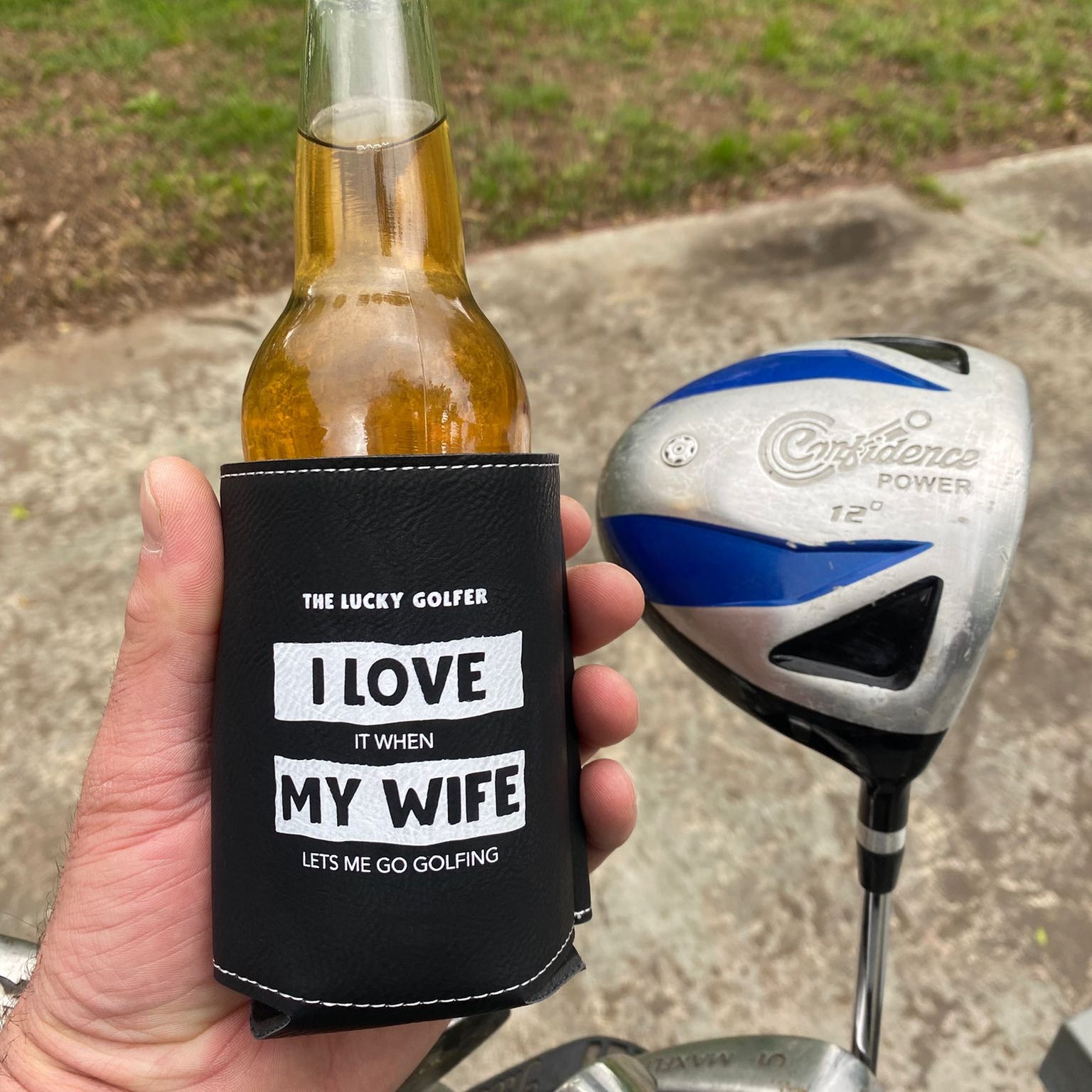 Hand holding a bottle with a can cooler labeled 'I Love My Wife' next to a golf club on a grassy background